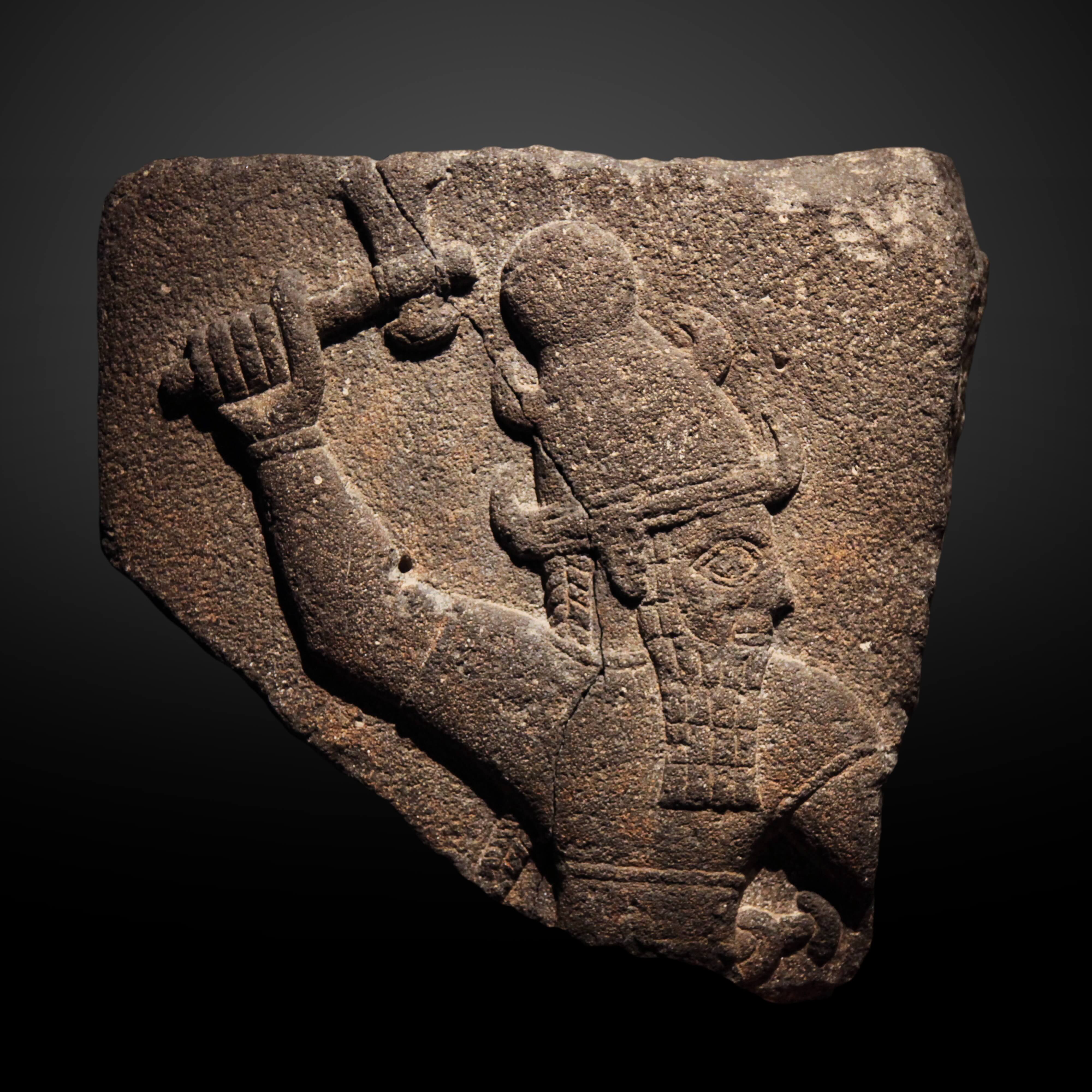 Photo of a basalt relief of the Hittite storm god Tarḫunt-. He is beared, wears an elaborate hat, and swings his hammer above his head.