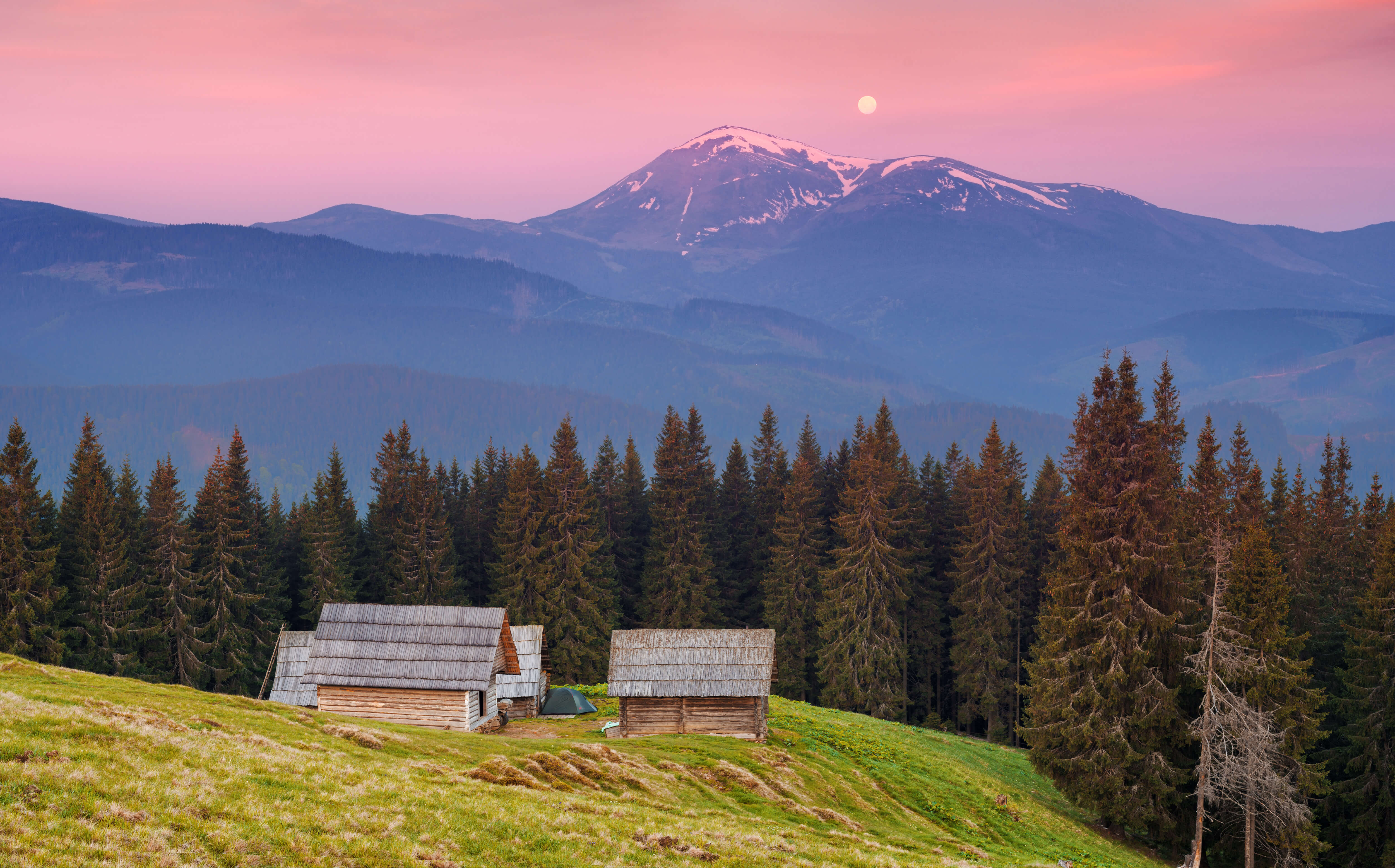 Pale sun in a pink morning lit sky behind two snow covered mountain tops. In the foreground, an idyllic wooden camping side on a green hill surrounded by pine forrest.